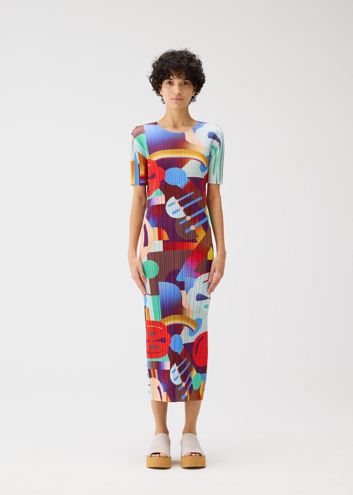 A person stands facing forward in the PLAYGROUND midi dress by PLEATS PLEASE ISSEY MIYAKE, featuring a colorful geometric pattern and short sleeves. They have short curly hair, a neutral expression, and wear light platform shoes against a plain white backdrop. view 4 of 6