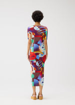 A person with short curly hair faces away, wearing the PLAYGROUND round-neck dress by PLEATS PLEASE ISSEY MIYAKE in a vibrant geometric pattern and tan platform sandals against a plain white background.