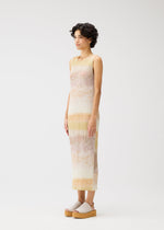 A person with short curly hair stands sideways, wearing the SEA BUBBLE sleeveless ankle-length pastel-striped beach dress by PLEATS PLEASE ISSEY MIYAKE and platform sandals. The background is plain white.