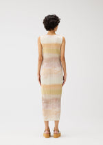 A person with short curly hair faces away, wearing the ankle-length ribbed SEA BUBBLE dress by PLEATS PLEASE ISSEY MIYAKE—sleeveless with horizontal pastel stripes and platform sandals—set against a plain white background.