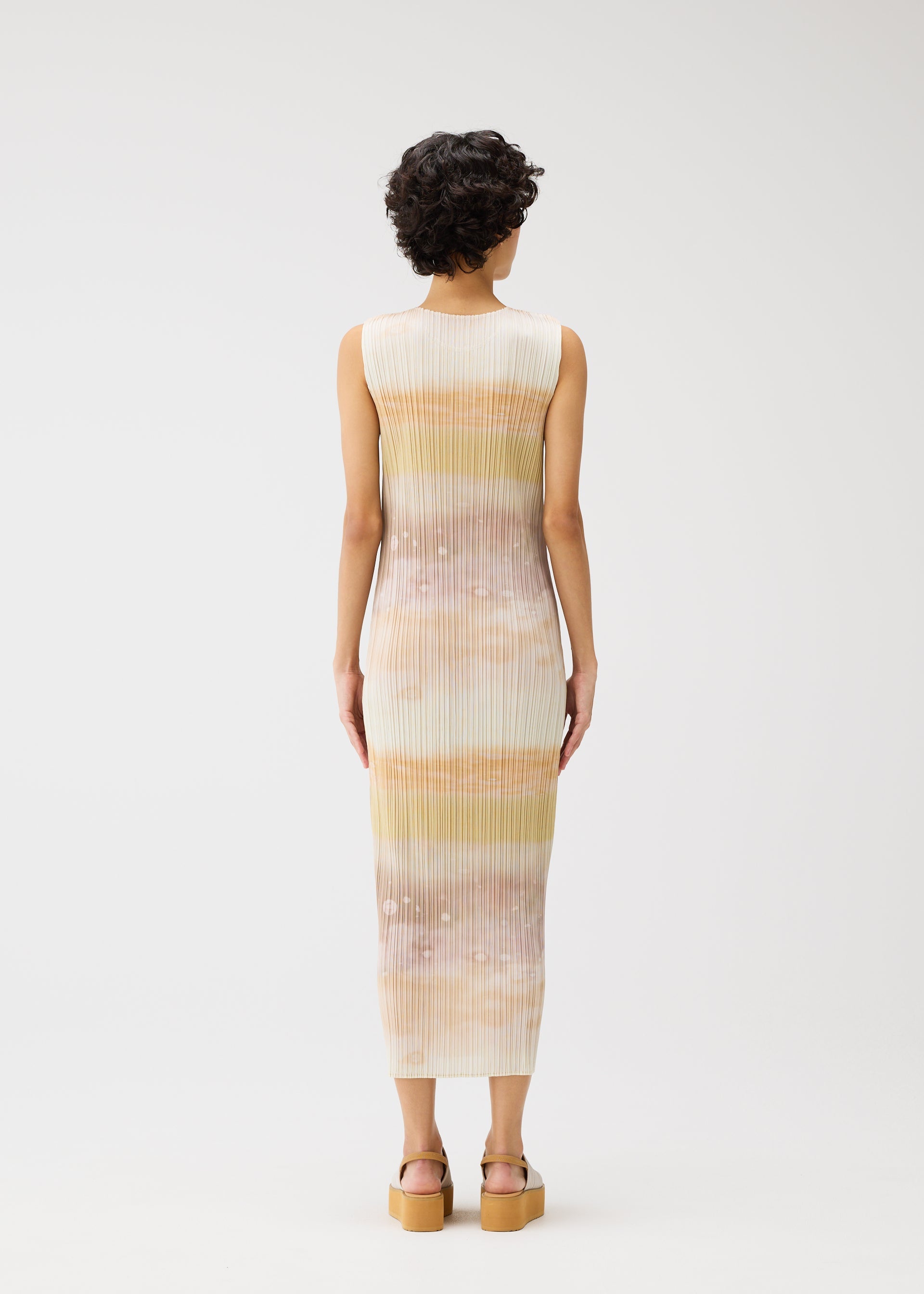 Zoomed view A person with short curly hair faces away, wearing the ankle-length ribbed SEA BUBBLE dress by PLEATS PLEASE ISSEY MIYAKE—sleeveless with horizontal pastel stripes and platform sandals—set against a plain white background.