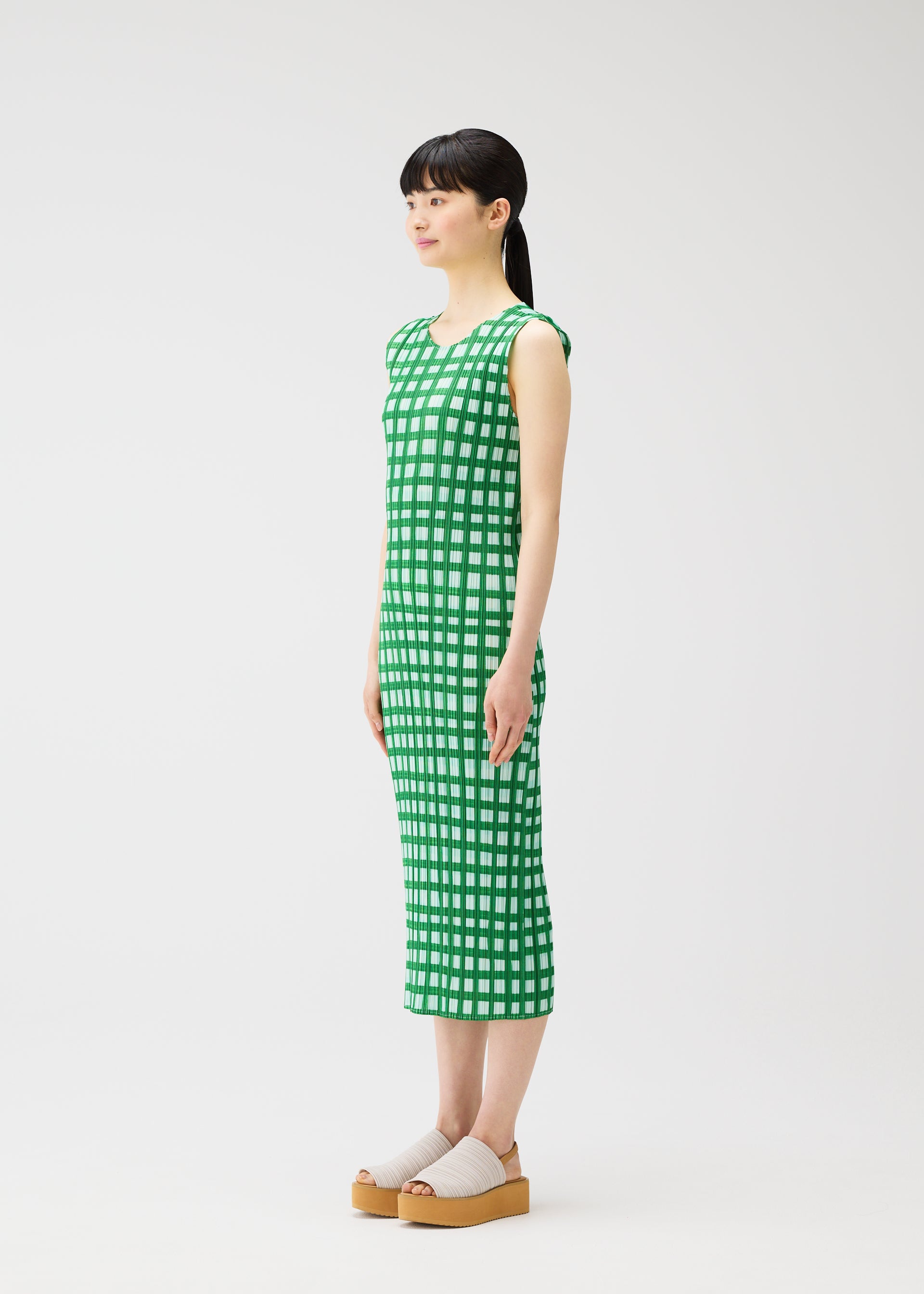 Zoomed view A woman stands in profile wearing the PLEATS PLEASE ISSEY MIYAKE BASKET green and white block check print sleeveless dress with beige platform sandals against a plain white background.