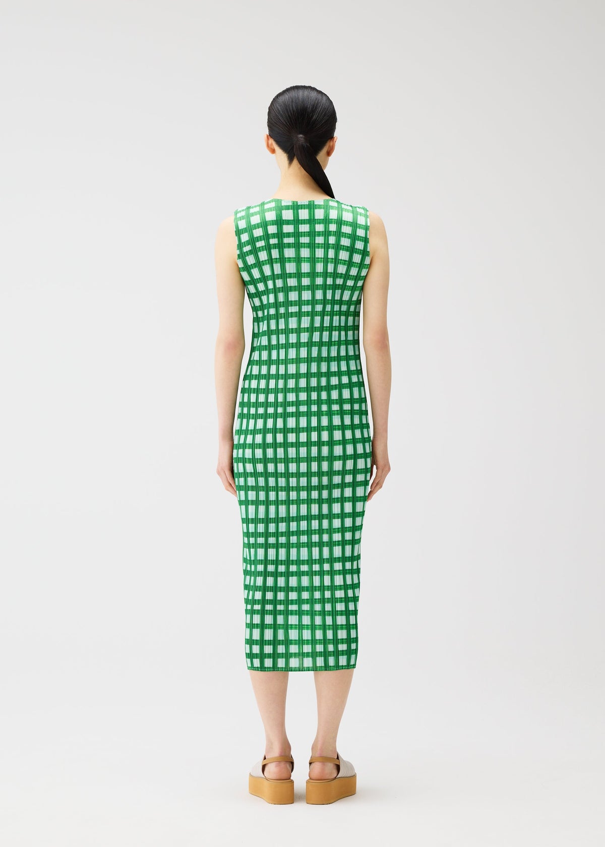 A woman with straight black hair in a low ponytail stands facing away, wearing the PLEATS PLEASE ISSEY MIYAKE BASKET green and white block check sleeveless midi dress and tan platform sandals against a plain white background. view 6 of 7