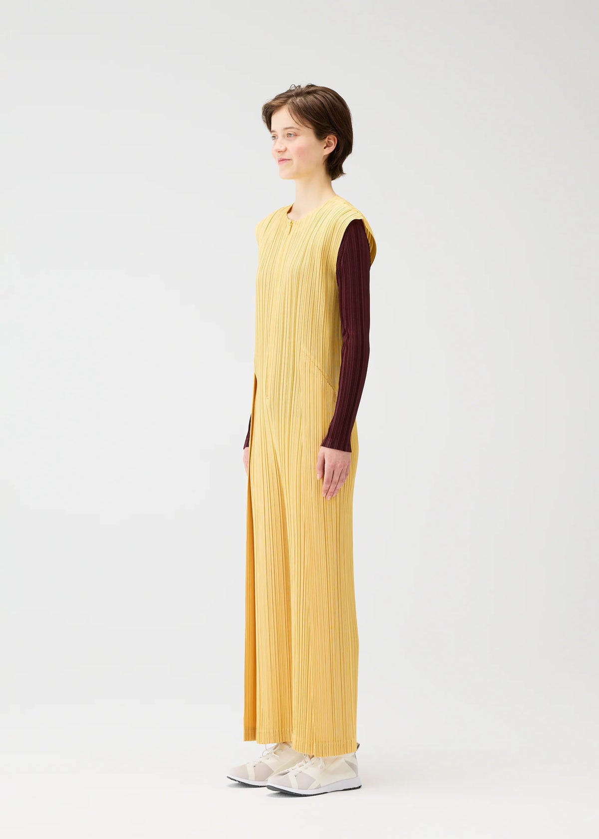A person stands in profile wearing PLEATS PLEASE ISSEY MIYAKE's THICKER BOTTOMS 1, a long sleeveless yellow pleated dress with pockets over a brown long-sleeve top and white shoes, set against a plain white background. view 8 of 10