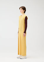 A person stands in profile wearing PLEATS PLEASE ISSEY MIYAKE's THICKER BOTTOMS 1, a long sleeveless yellow pleated dress with pockets over a brown long-sleeve top and white shoes, set against a plain white background.