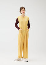 A person stands against a plain white background, wearing PLEATS PLEASE ISSEY MIYAKE's THICKER BOTTOMS 1—a zippered yellow sleeveless jumpsuit with pockets—over a maroon long-sleeve shirt and white sneakers, hands in pockets, looking relaxed.