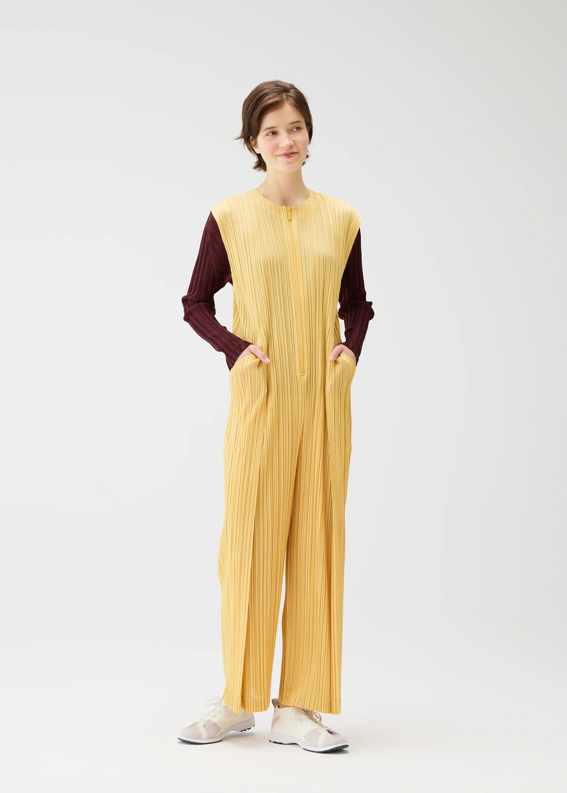 Zoomed view A person stands against a plain white background, wearing PLEATS PLEASE ISSEY MIYAKE's THICKER BOTTOMS 1—a zippered yellow sleeveless jumpsuit with pockets—over a maroon long-sleeve shirt and white sneakers, hands in pockets, looking relaxed.