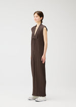 A person stands in profile against a plain white background, wearing LOOP BOTTOMS by PLEATS PLEASE ISSEY MIYAKE—a long, sleeveless dark brown pleated dress with a deep V-neckline—paired with white socks and translucent shoes.