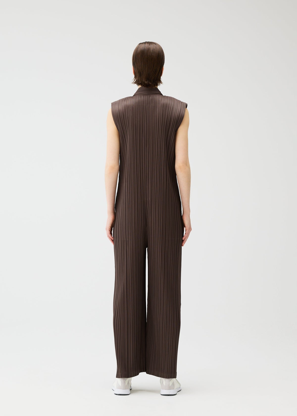 A person faces away, wearing LOOP BOTTOMS by PLEATS PLEASE ISSEY MIYAKE—a sleeveless, loose-fitting, dark brown pleated jumpsuit made from recycled polyester—with white shoes, set against a plain light background. view 7 of 8