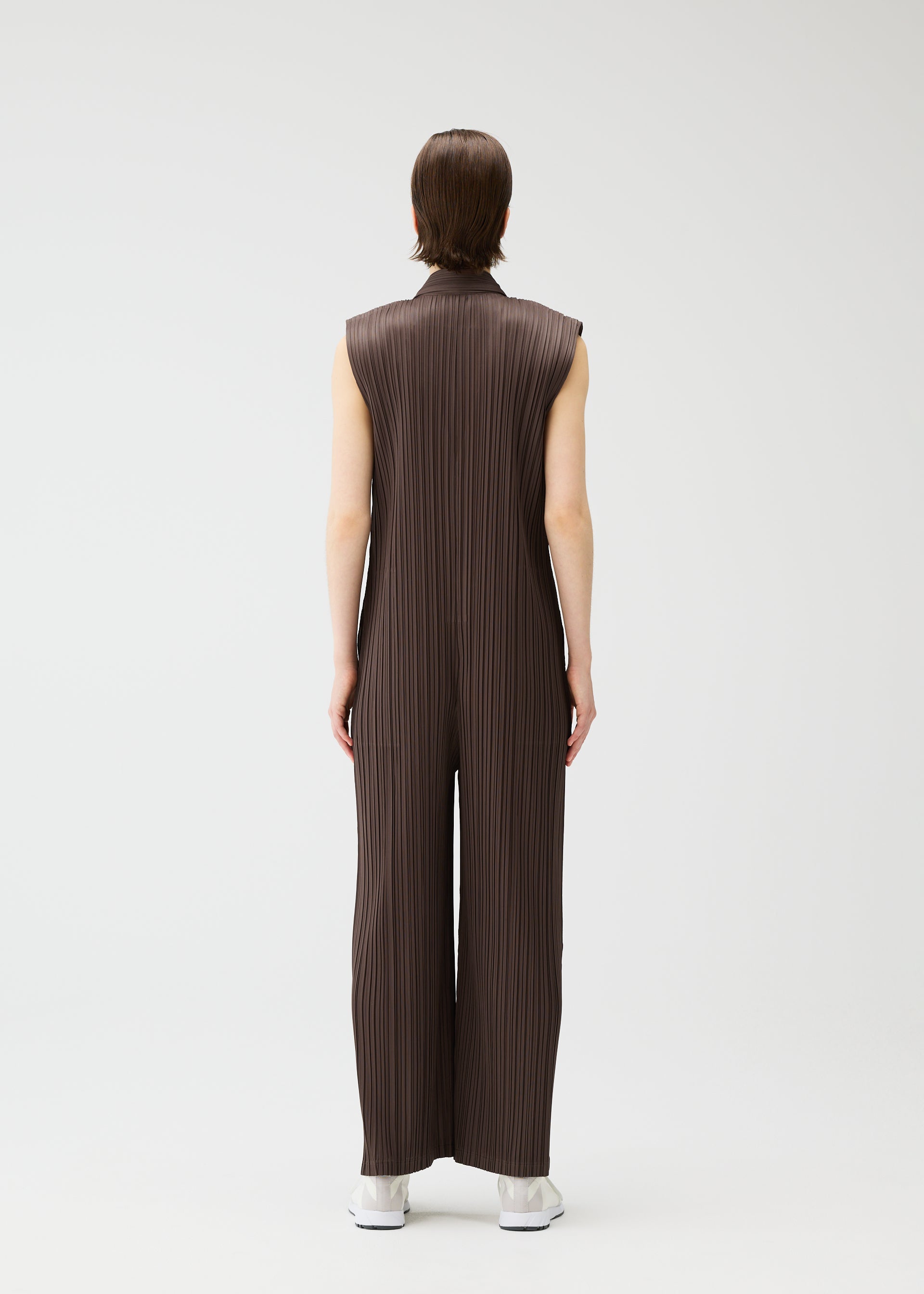 Zoomed view A person faces away, wearing LOOP BOTTOMS by PLEATS PLEASE ISSEY MIYAKE—a sleeveless, loose-fitting, dark brown pleated jumpsuit made from recycled polyester—with white shoes, set against a plain light background.