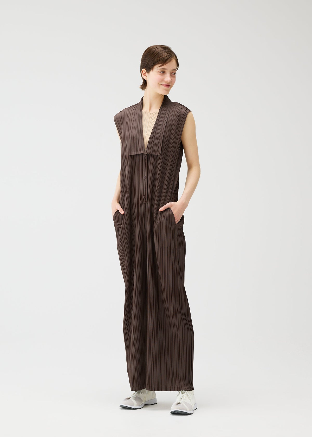 A person smiles and looks to the side against a white background, wearing PLEATS PLEASE ISSEY MIYAKE LOOP BOTTOMS—a sleeveless, loose brown jumpsuit with button closures, wide legs, side pockets—and white sneakers. view 8 of 8