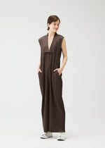 A person smiles and looks to the side against a white background, wearing PLEATS PLEASE ISSEY MIYAKE LOOP BOTTOMS—a sleeveless, loose brown jumpsuit with button closures, wide legs, side pockets—and white sneakers.