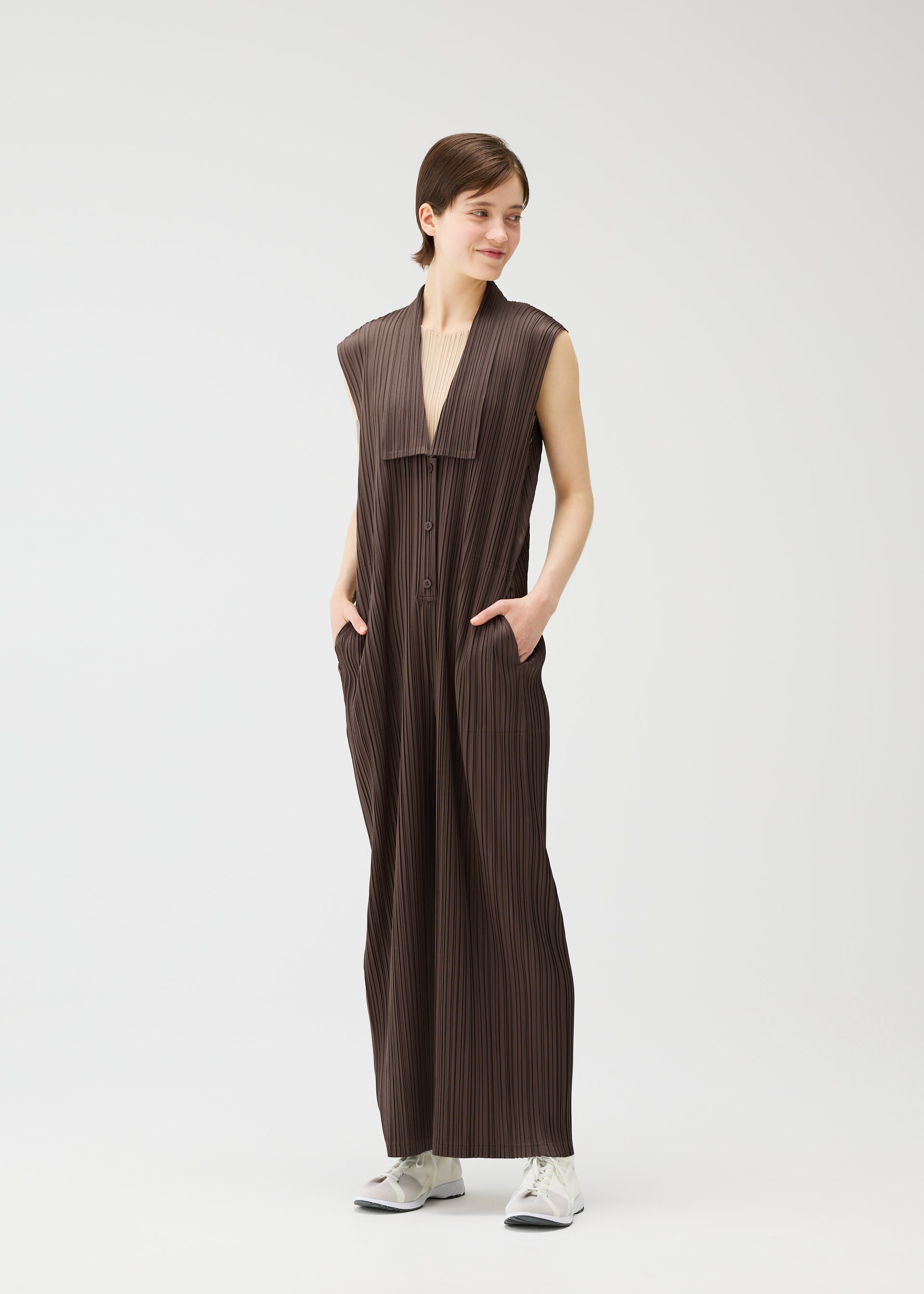 Zoomed view A person smiles and looks to the side against a white background, wearing PLEATS PLEASE ISSEY MIYAKE LOOP BOTTOMS—a sleeveless, loose brown jumpsuit with button closures, wide legs, side pockets—and white sneakers.