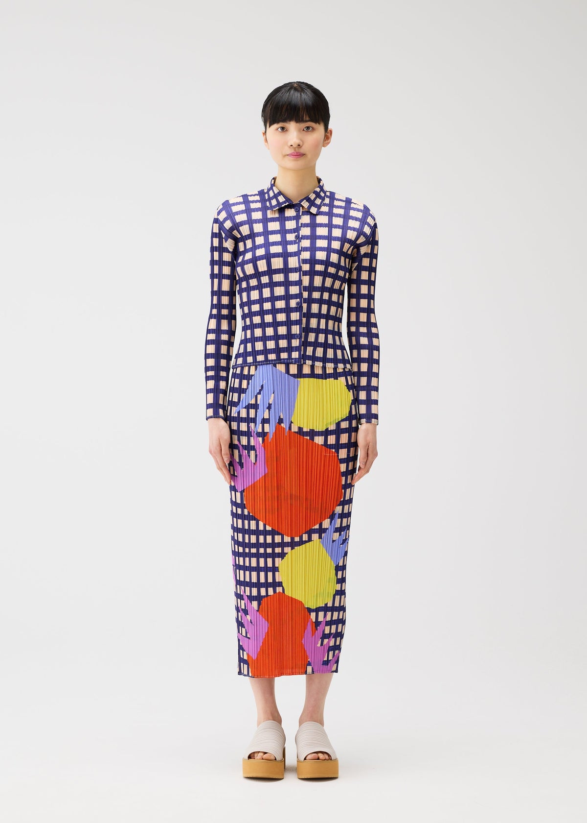 A woman with short dark hair stands facing forward, holding the BASKET by PLEATS PLEASE ISSEY MIYAKE. She wears a long-sleeved, ankle-length dress in blue and white block checks with abstract shapes, beige platform shoes, against a white background. view 4 of 6