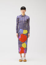 A woman with short dark hair stands facing forward, holding the BASKET by PLEATS PLEASE ISSEY MIYAKE. She wears a long-sleeved, ankle-length dress in blue and white block checks with abstract shapes, beige platform shoes, against a white background.