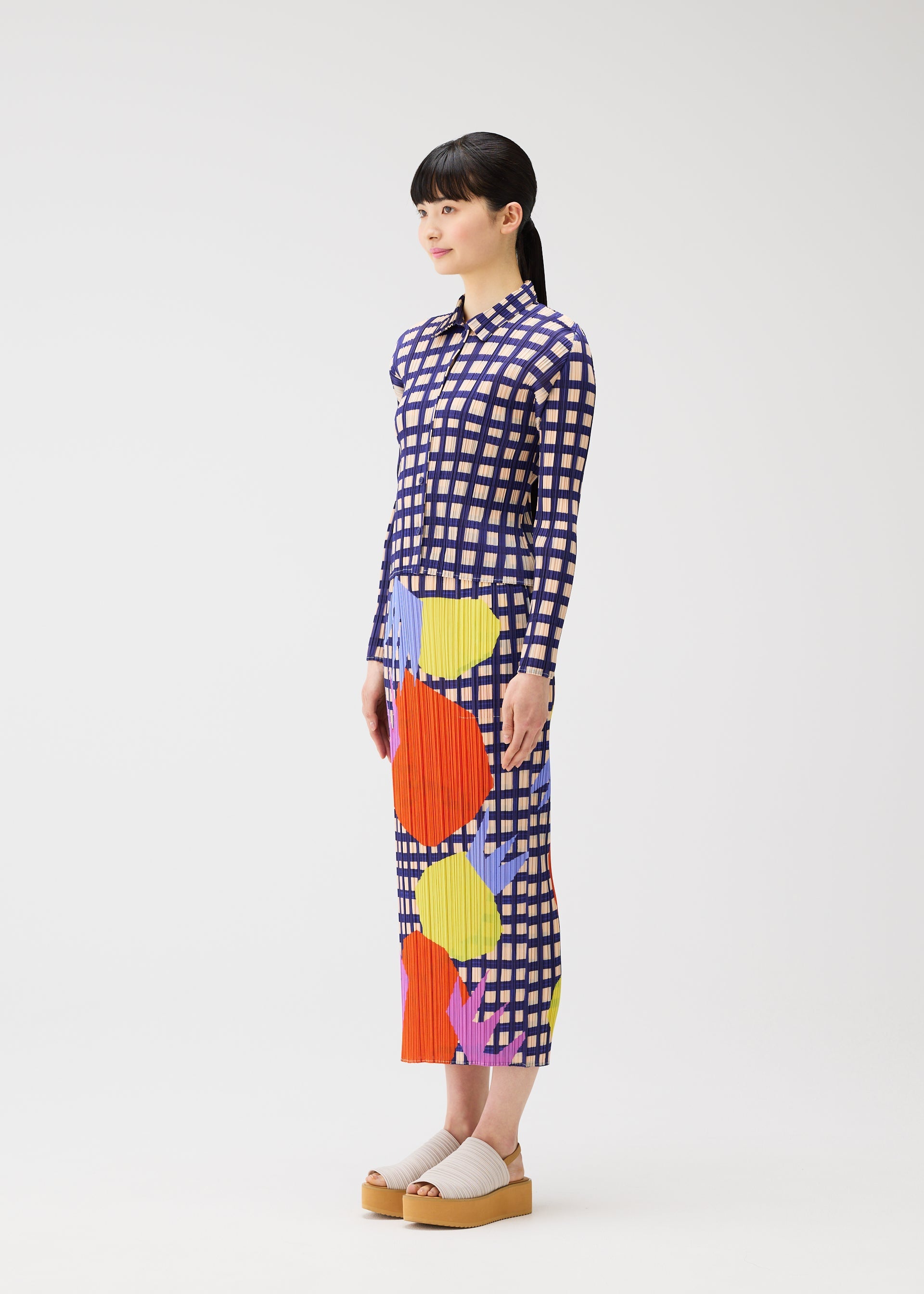 Zoomed view A woman with straight black hair and bangs models the BASKET dress by PLEATS PLEASE ISSEY MIYAKE, a long-sleeve, ankle-length design with colorful block check prints. She pairs it with beige platform sandals against a plain white background.