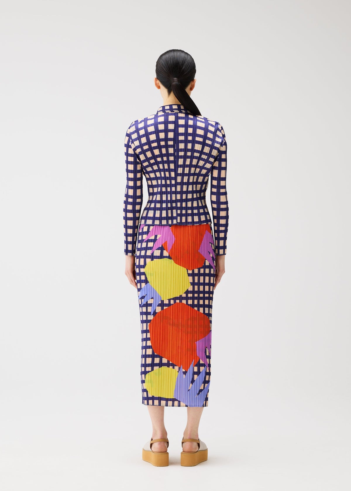 A woman stands facing away, wearing the BASKET ankle-length dress by PLEATS PLEASE ISSEY MIYAKE, featuring a block check print with large red, yellow, and purple shapes. Her dark hair is tied back and she wears platform sandals. view 6 of 6