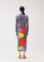A woman stands facing away, wearing the BASKET ankle-length dress by PLEATS PLEASE ISSEY MIYAKE, featuring a block check print with large red, yellow, and purple shapes. Her dark hair is tied back and she wears platform sandals.