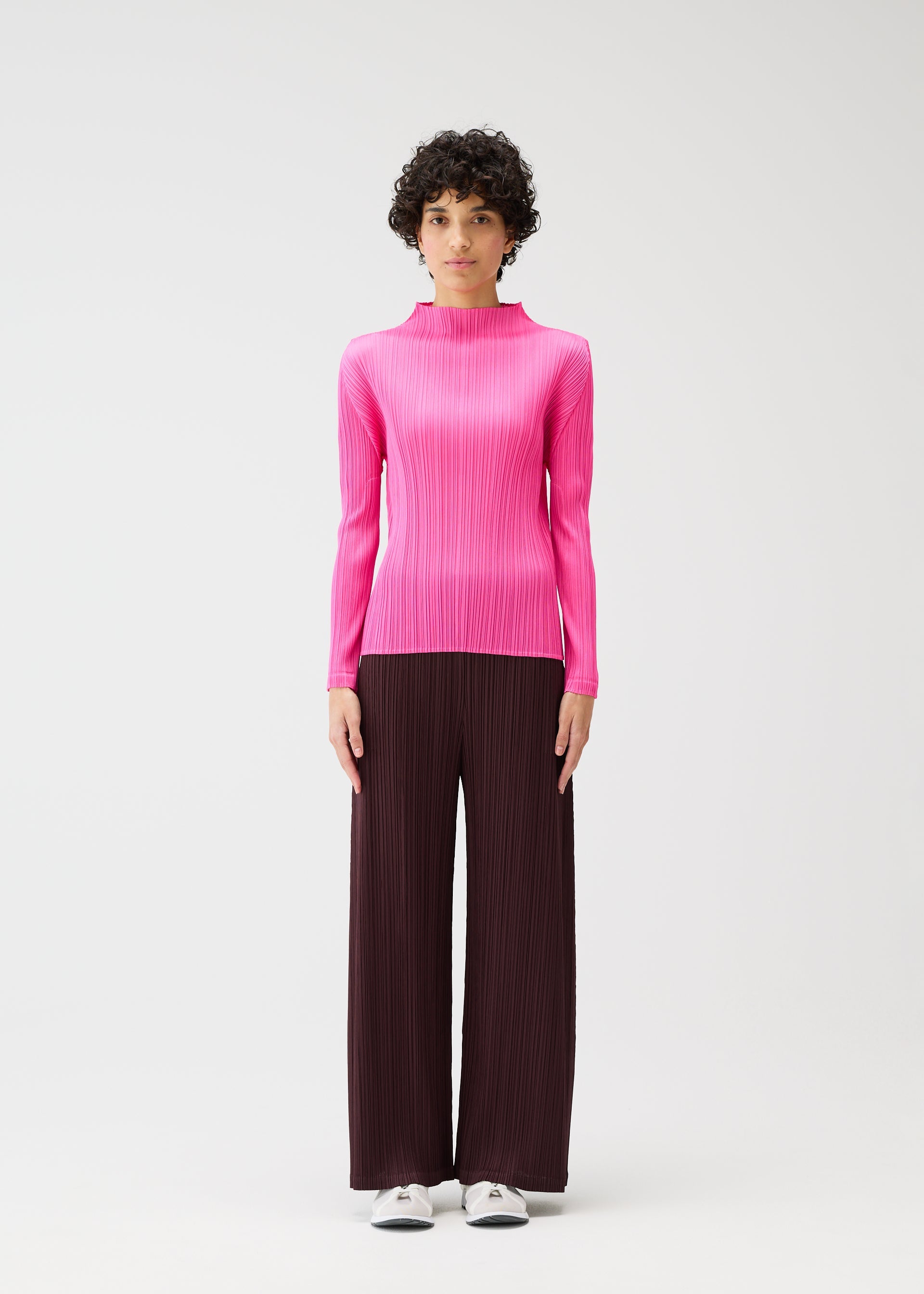 Zoomed view A person with short curly hair faces forward, wearing the bright pink pleated top and dark brown pleated pants from PLEATS PLEASE ISSEY MIYAKE's MONTHLY COLORS : JANUARY collection, with white sneakers, against a plain background inspired by townscapes.