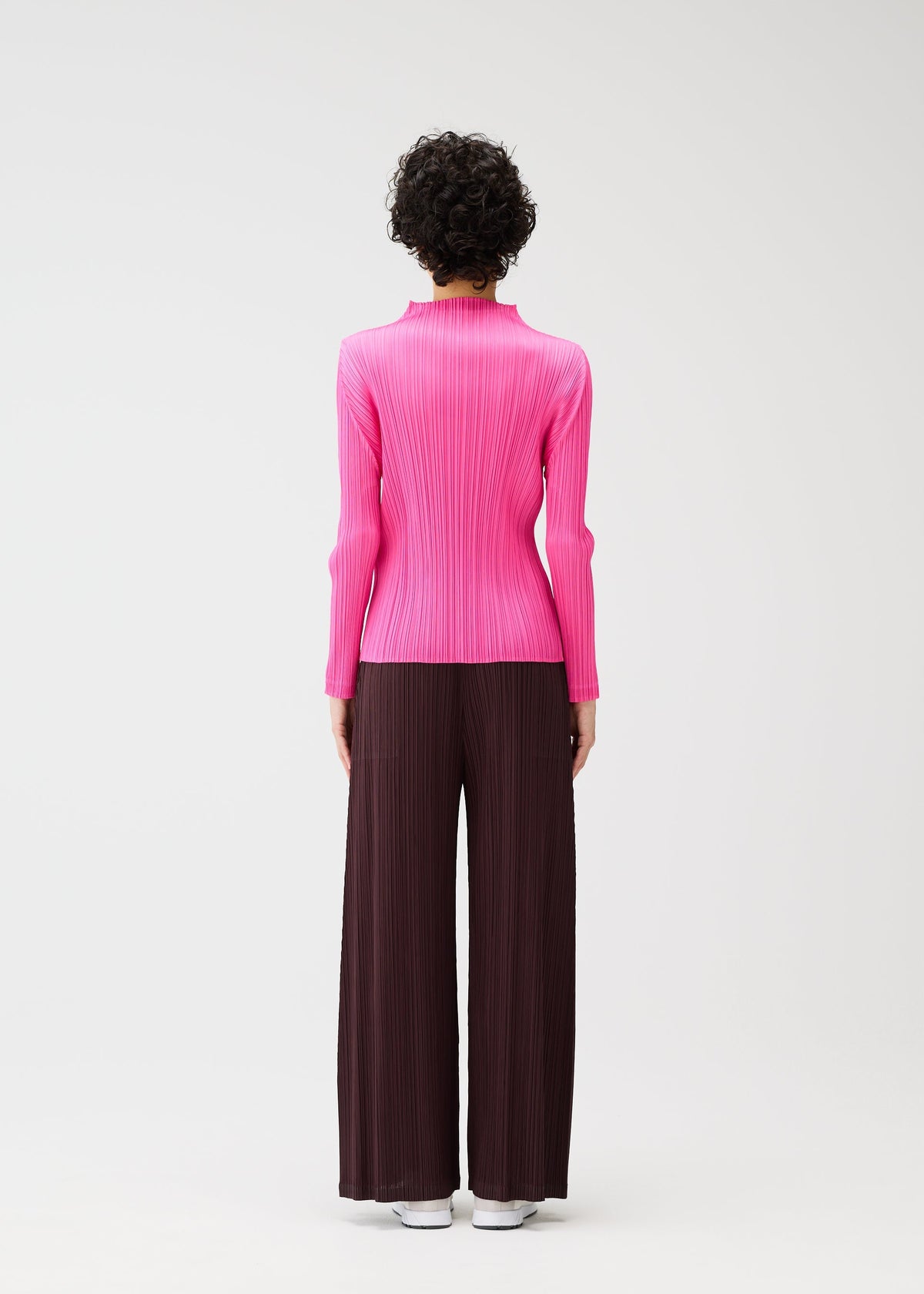 A person faces away against a white background, wearing the PLEATS PLEASE ISSEY MIYAKE "MONTHLY COLORS : JANUARY" bright pink long-sleeved top and loose, dark brown pleated pants, paired with white shoes. view 9 of 10