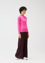 A person with short curly hair smiles in a bright pink long-sleeved MONTHLY COLORS : JANUARY top and wide-legged dark brown pants by PLEATS PLEASE ISSEY MIYAKE, paired with white sneakers against a plain white background.