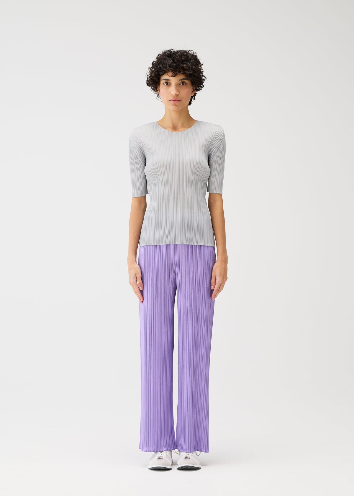 A person with short curly hair models the PLEATS PLEASE ISSEY MIYAKE MONTHLY COLORS: APRIL collection, wearing a light gray top and pleated lavender pants with white shoes against a plain white background. view 7 of 10