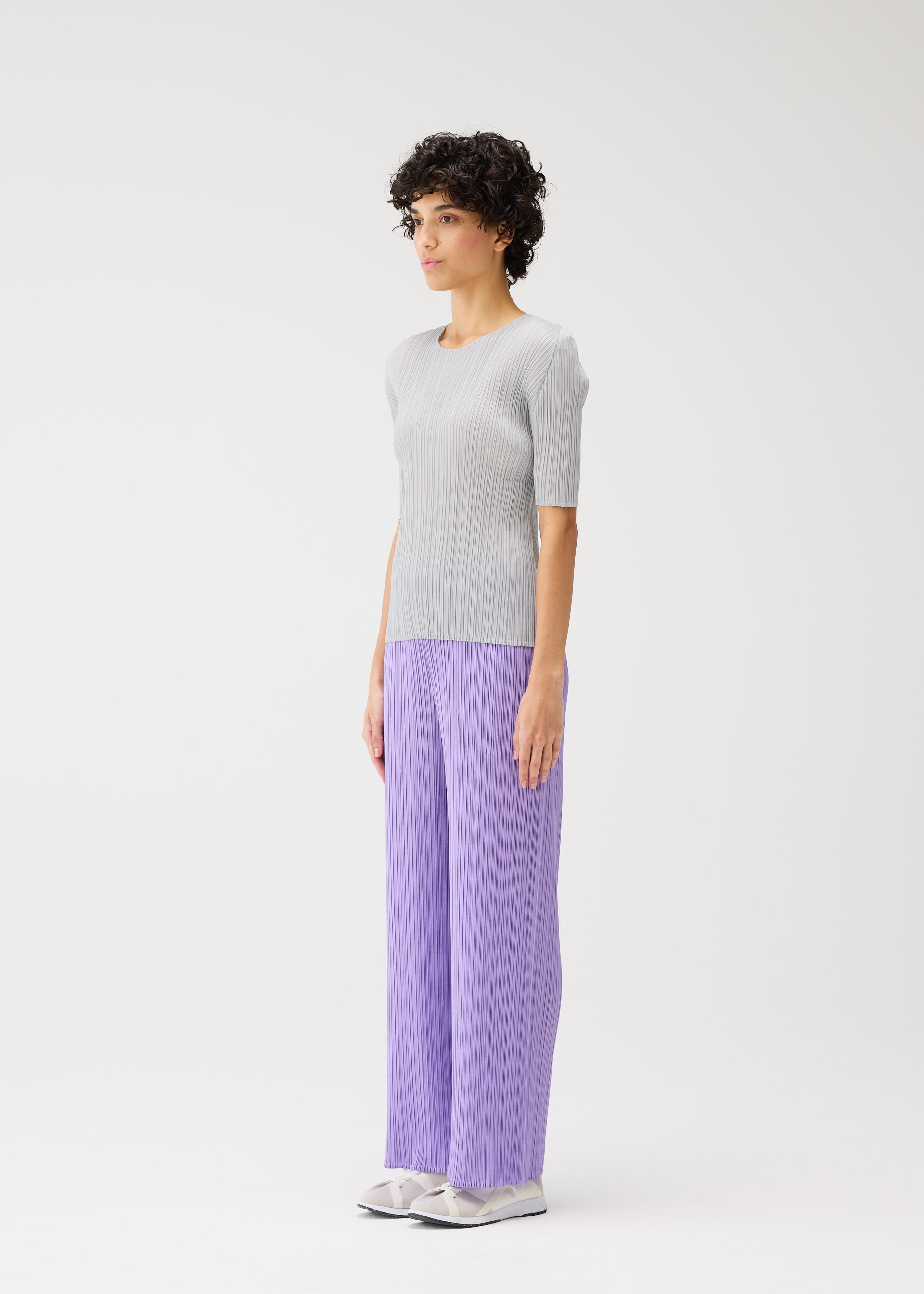 Zoomed view A person with short curly hair stands in profile, wearing the MONTHLY COLORS : APRIL wide-legged lavender pants by PLEATS PLEASE ISSEY MIYAKE, a round-neck top, and white shoes against a plain white background.