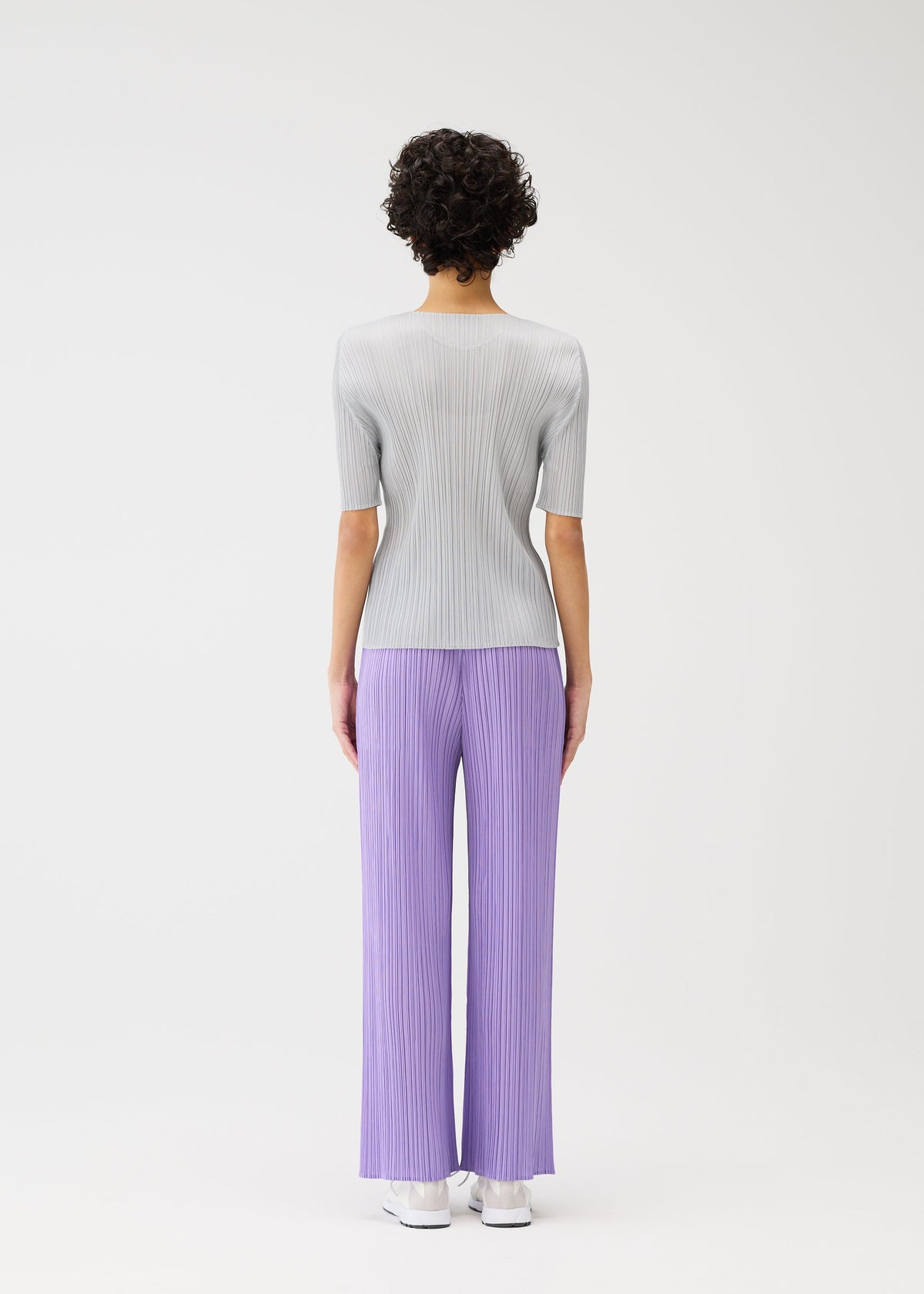 A person with short curly hair faces away, modeling PLEATS PLEASE ISSEY MIYAKE's MONTHLY COLORS : APRIL—light gray ribbed round-neck top and loose, pleated lavender pants with white shoes—against a plain white backdrop. view 9 of 10