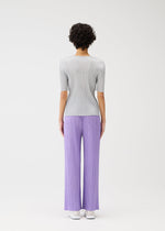 A person with short curly hair faces away, modeling PLEATS PLEASE ISSEY MIYAKE's MONTHLY COLORS : APRIL—light gray ribbed round-neck top and loose, pleated lavender pants with white shoes—against a plain white backdrop.