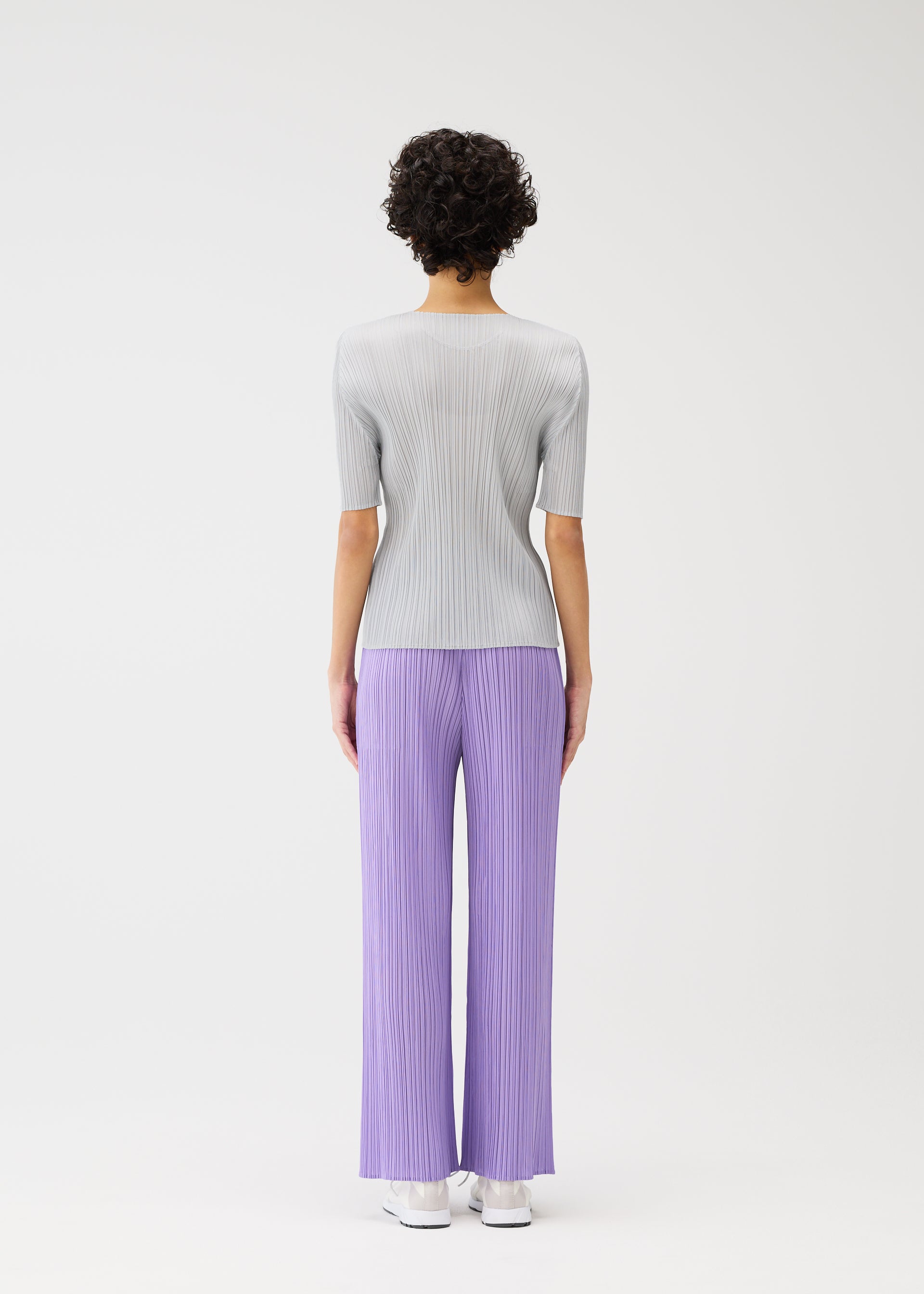 Zoomed view A person with short curly hair faces away, modeling PLEATS PLEASE ISSEY MIYAKE's MONTHLY COLORS : APRIL—light gray ribbed round-neck top and loose, pleated lavender pants with white shoes—against a plain white backdrop.