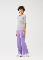 A person with short curly hair models the MONTHLY COLORS : APRIL collection from PLEATS PLEASE ISSEY MIYAKE, featuring a light gray ribbed top and loose lavender pleated pants with white sneakers, set against a plain white background.