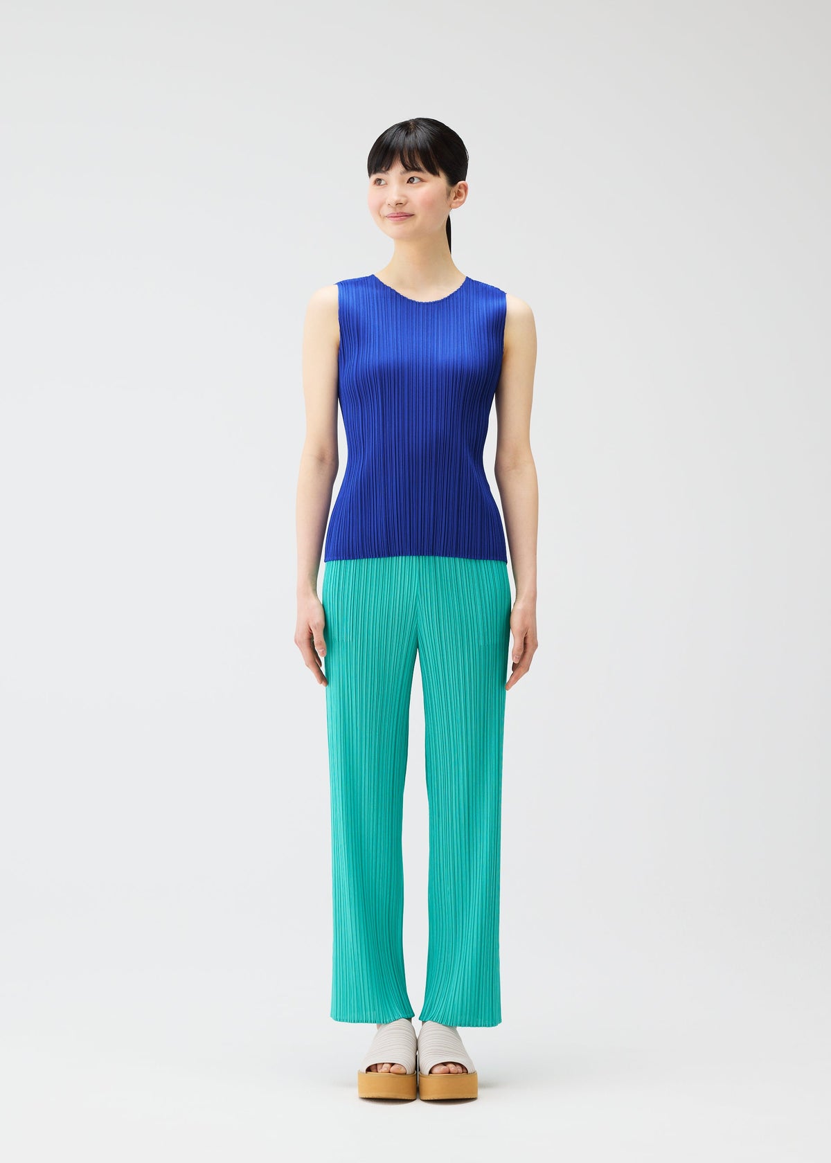 A woman models the PLEATS PLEASE ISSEY MIYAKE MONTHLY COLORS : MAY collection—sporting a blue sleeveless ribbed tank top and teal ribbed pants with beige platform sandals, her straight dark hair and smile embody effortless summer style. view 7 of 10