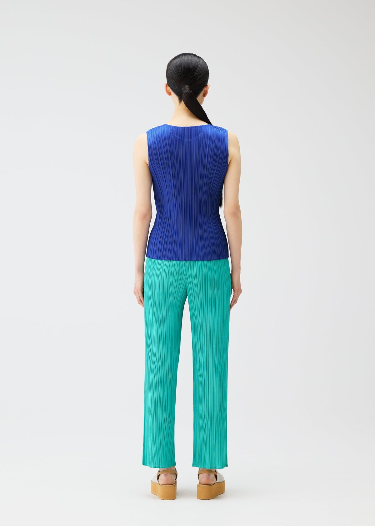A person faces away against a white background, wearing PLEATS PLEASE ISSEY MIYAKE's MONTHLY COLORS : MAY—a sleeveless blue tank top with subtle gradations, teal pleated pants, and tan platform sandals. Their dark hair is tied back for a chic summer look. view 9 of 10