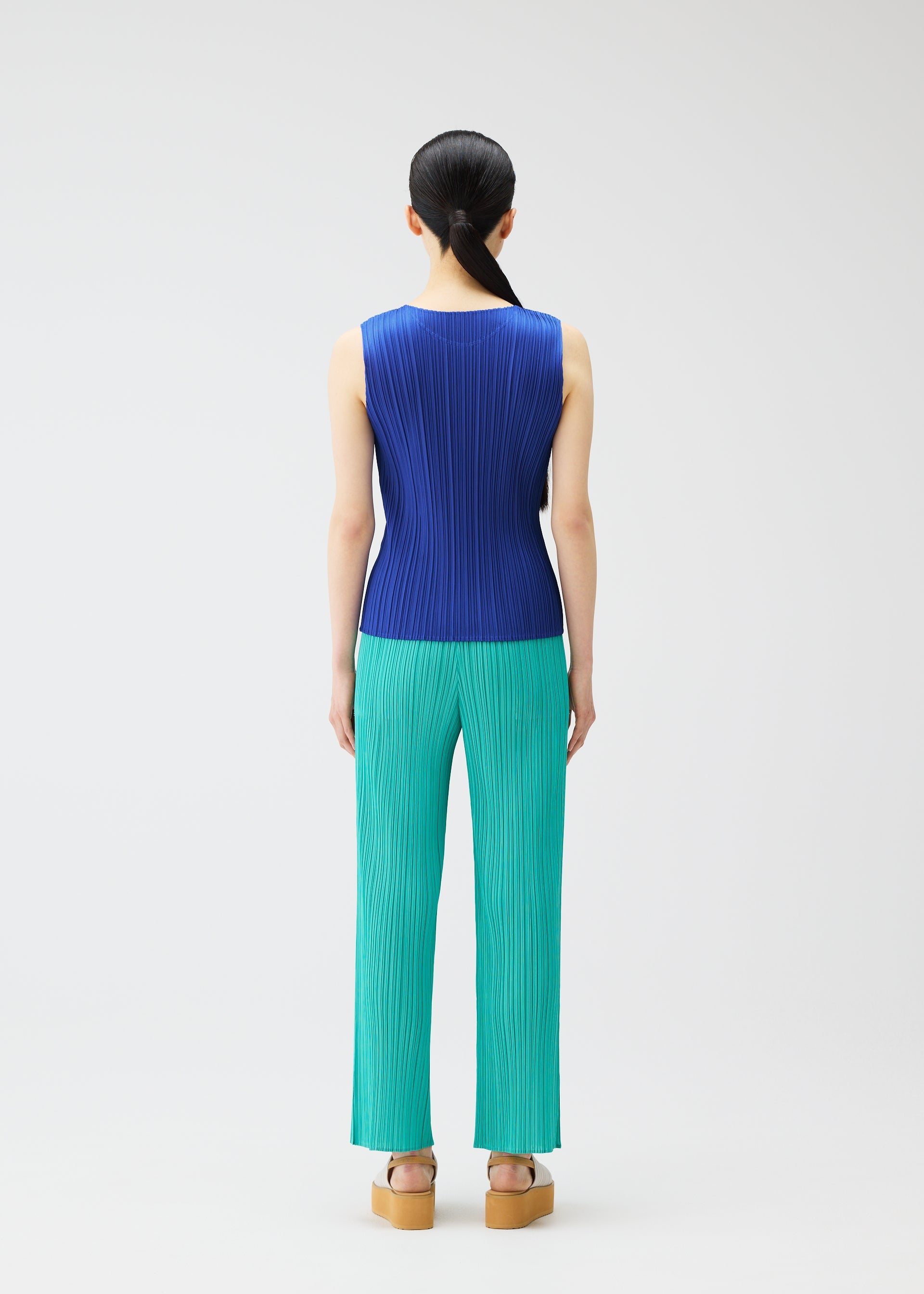 Zoomed view A person faces away against a white background, wearing PLEATS PLEASE ISSEY MIYAKE's MONTHLY COLORS : MAY—a sleeveless blue tank top with subtle gradations, teal pleated pants, and tan platform sandals. Their dark hair is tied back for a chic summer look.