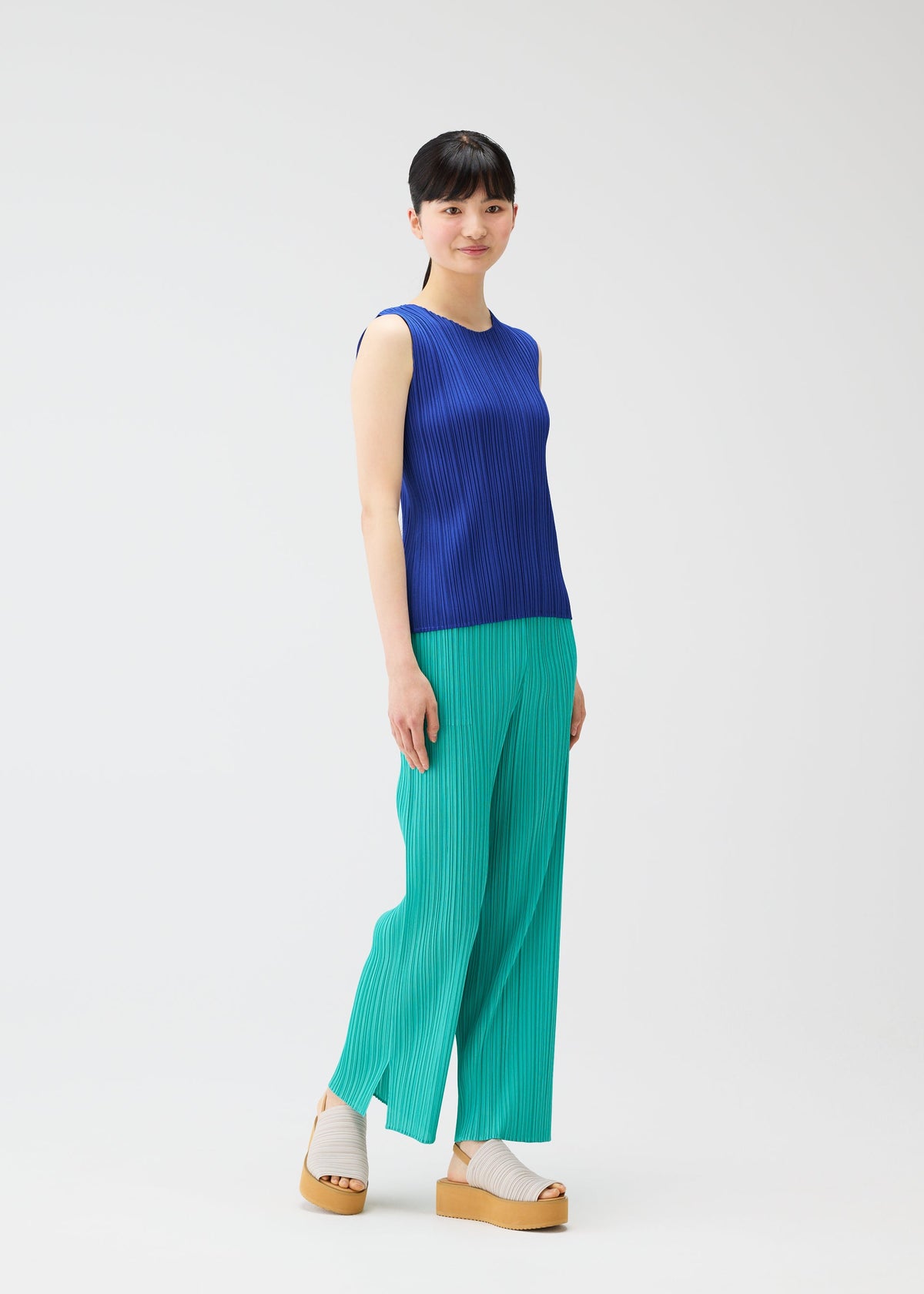 A woman models the MONTHLY COLORS : MAY collection by PLEATS PLEASE ISSEY MIYAKE, wearing a sleeveless ribbed blue tank, green ribbed pants, and beige platform sandals against a plain white background, her hair tied back and smiling softly. view 10 of 10