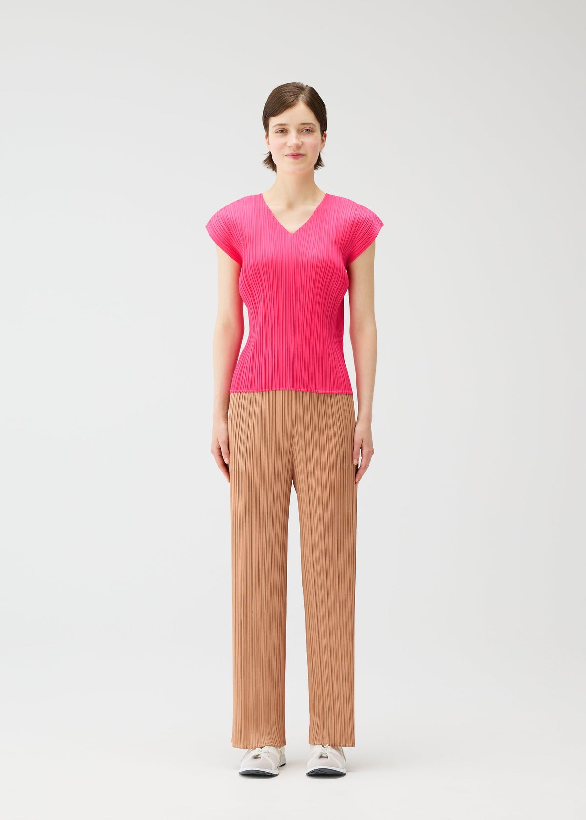 A woman with short brown hair and a neutral expression wears the MONTHLY COLORS : JUNE bright pink pleated top and beige wide-legged pleated pants from PLEATS PLEASE ISSEY MIYAKE, styled with white sneakers against a plain white background. view 7 of 10