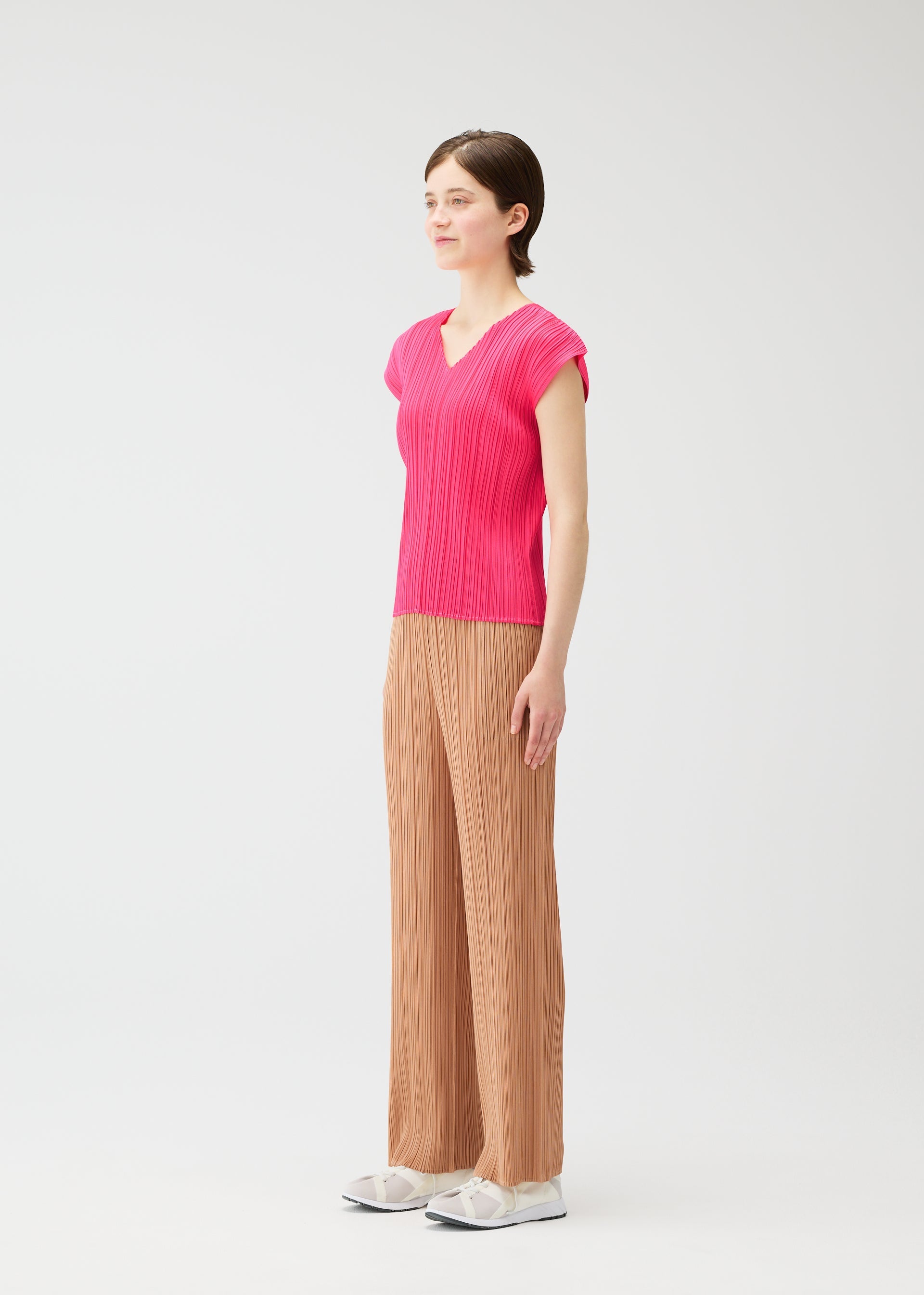 Zoomed view A person models the PLEATS PLEASE ISSEY MIYAKE MONTHLY COLORS : JUNE collection in a bright pink V-neck top and loose tan pleated pants with white shoes, standing in a light studio with a plain white background, facing slightly left.