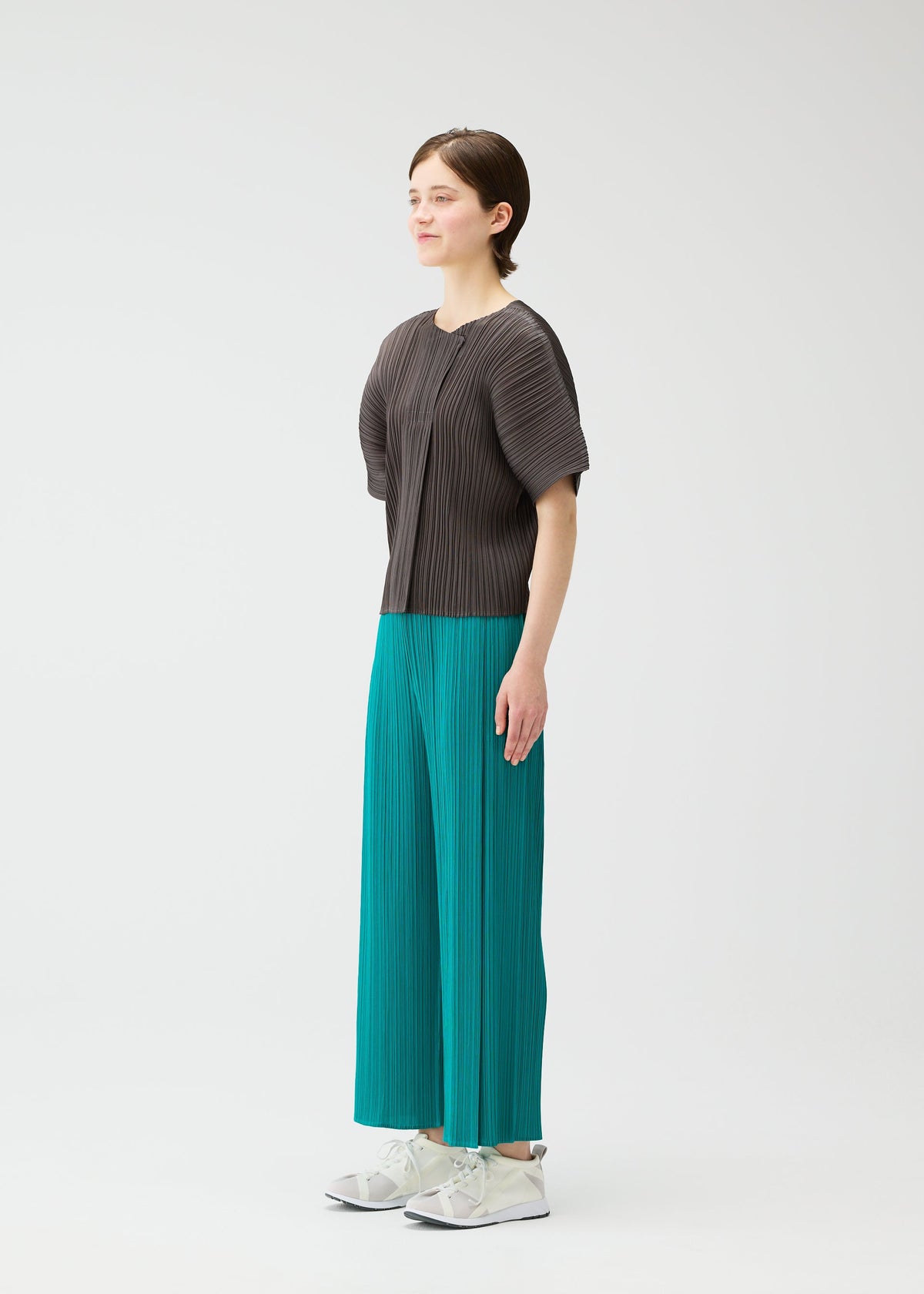 A person stands against a plain white background, wearing PLEATS PLEASE ISSEY MIYAKE's MONTHLY COLORS: JUNE collection—a dark gray pleated top and bright teal pleated wide-leg pants—with white sneakers, facing slightly left with arms relaxed. view 8 of 11