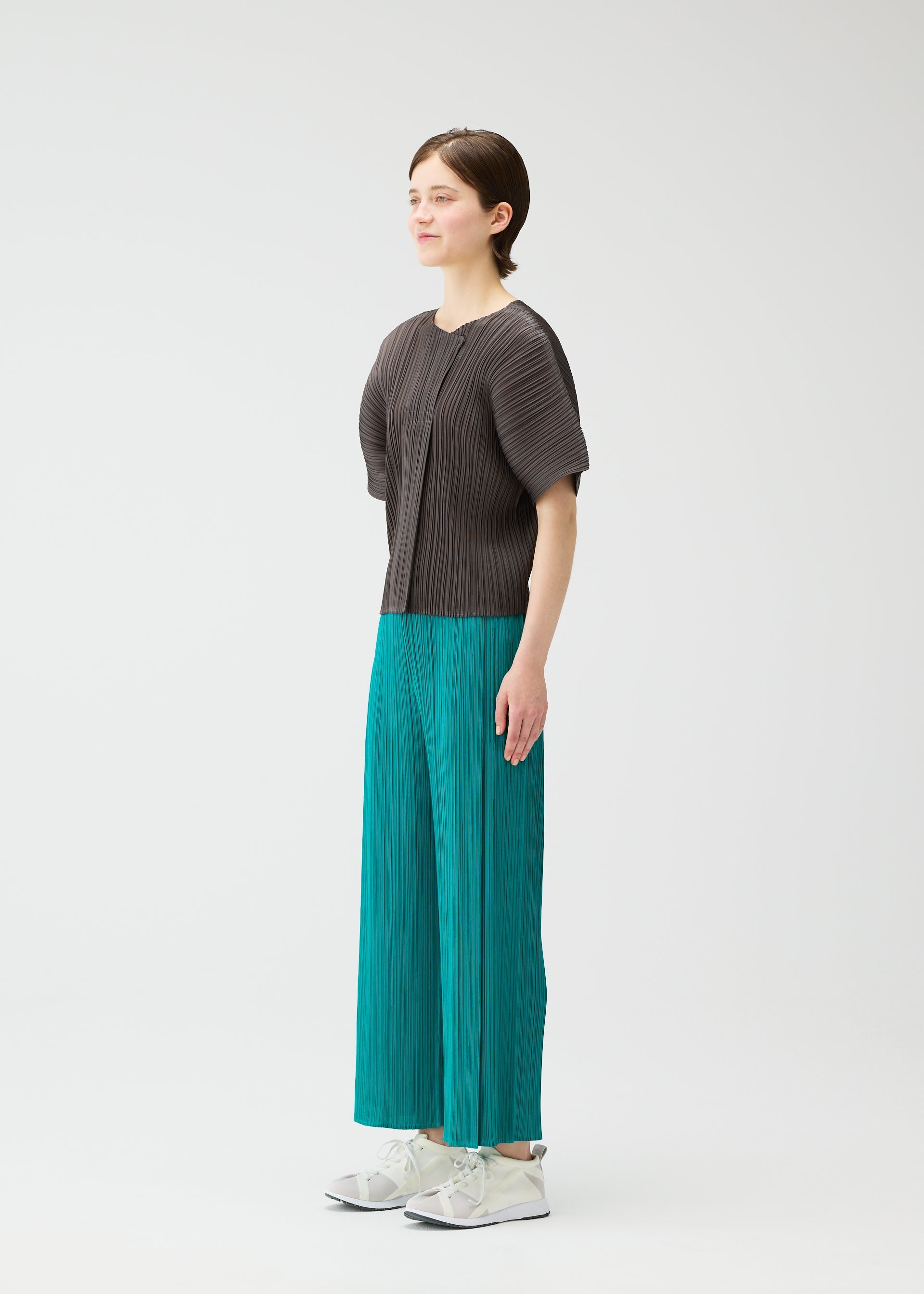 Zoomed view A person stands against a plain white background, wearing PLEATS PLEASE ISSEY MIYAKE's MONTHLY COLORS: JUNE collection—a dark gray pleated top and bright teal pleated wide-leg pants—with white sneakers, facing slightly left with arms relaxed.