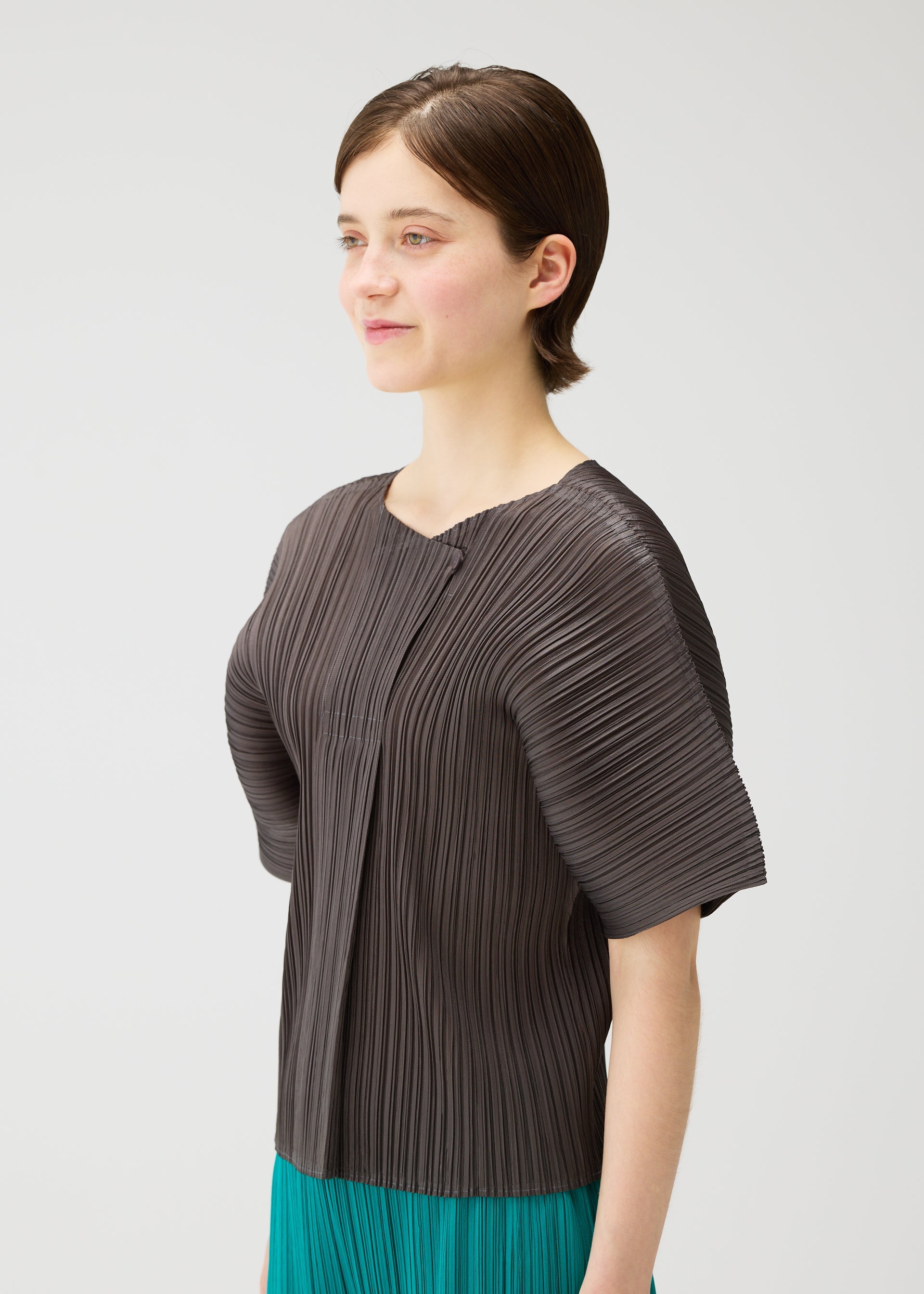 Zoomed view A young woman with short brown hair models the PLEATS PLEASE ISSEY MIYAKE MONTHLY COLORS : JUNE collection, wearing a dark, reversible wide-sleeved top and a green pleated skirt, standing against a plain light background and smiling softly.
