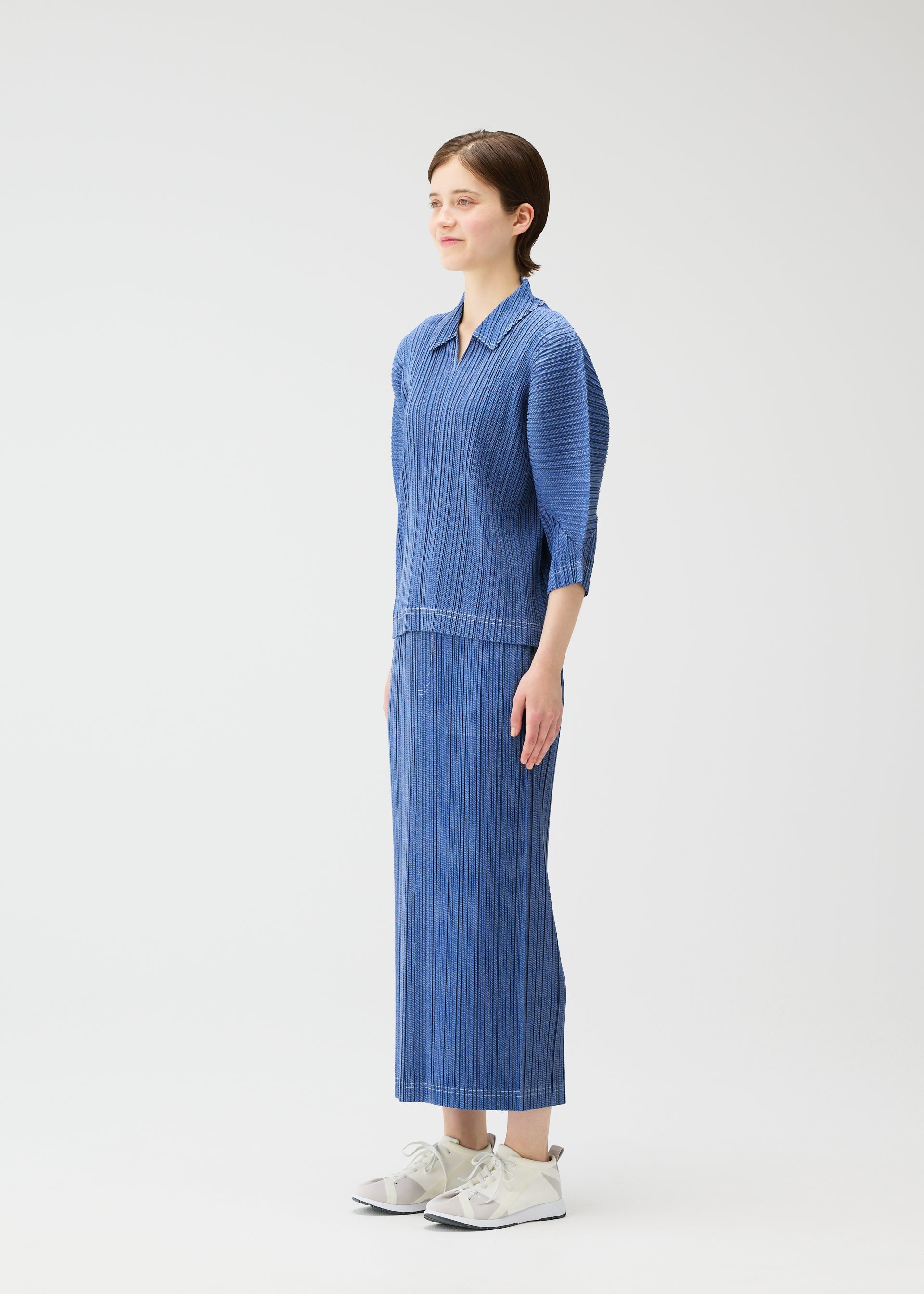 Zoomed view A person stands sideways against a plain white background, wearing the COTTON DENIM long-sleeve top and ankle-length skirt by PLEATS PLEASE ISSEY MIYAKE, paired with white sneakers.