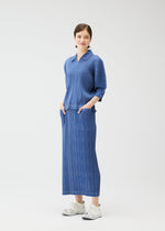 A woman poses against a white backdrop in the PLEATS PLEASE ISSEY MIYAKE COTTON DENIM set—a blue long-sleeve top and matching skirt in soft cotton blend, with hands in her pockets, finished with white sneakers.