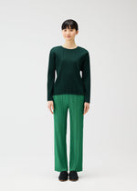 A woman faces forward against a white background, wearing PLEATS PLEASE ISSEY MIYAKE’s RAMIE PLEATS: a dark green long-sleeve round-neck ramie top and light green pleated pants, paired with black shoes.