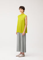 A person with short dark hair smiles softly against a plain background, dressed in the MELLOW PLEATS sleeveless high-neck yellow top and wide-leg gray pants by PLEATS PLEASE ISSEY MIYAKE, paired with light slip-on shoes.