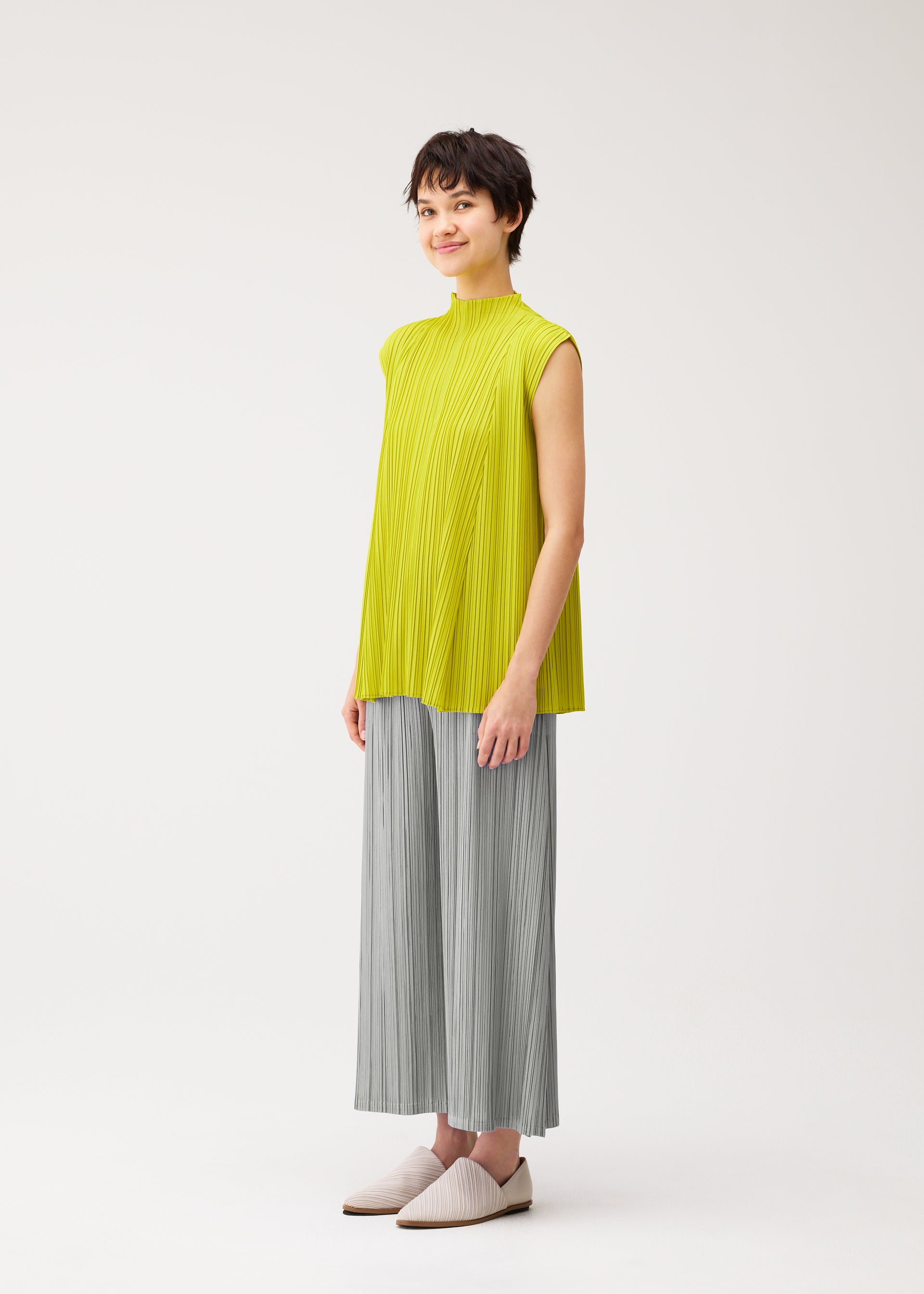 Zoomed view A person with short dark hair smiles softly against a plain background, dressed in the MELLOW PLEATS sleeveless high-neck yellow top and wide-leg gray pants by PLEATS PLEASE ISSEY MIYAKE, paired with light slip-on shoes.