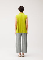 A person with short dark hair stands facing away, wearing the MELLOW PLEATS set by PLEATS PLEASE ISSEY MIYAKE—a sleeveless yellow-green high-neck top and wide-leg light grey pleated pants—paired with white slip-on shoes against a plain white background.