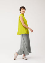 A woman with short dark hair smiles while walking in the MELLOW PLEATS top and loose gray pleated pants by PLEATS PLEASE ISSEY MIYAKE, paired with white slip-ons, against a plain white background.