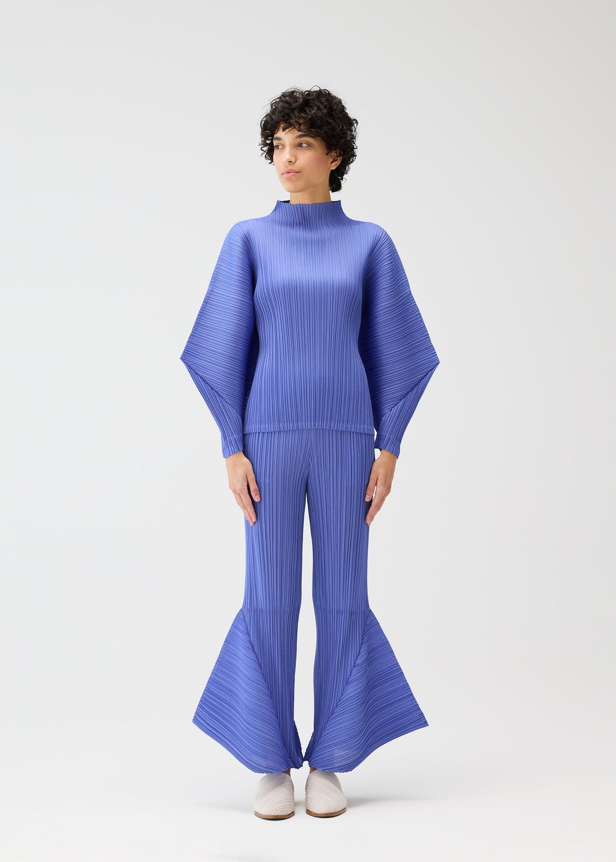 A person wearing the SKYLINE set by PLEATS PLEASE ISSEY MIYAKE—featuring a high-neck top with dolman sleeves and blue flared pleated pants—stands against a white background, styled with short curly hair and white shoes, gazing to the side. view 5 of 8