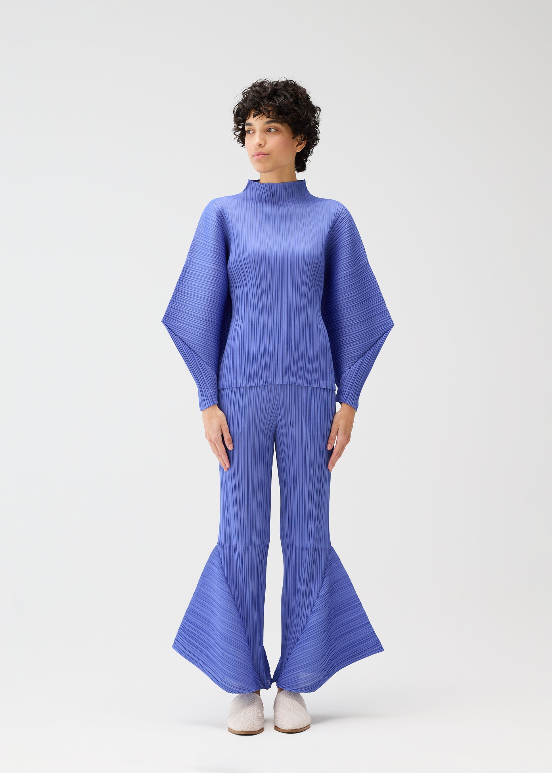 Zoomed view A person wearing the SKYLINE set by PLEATS PLEASE ISSEY MIYAKE—featuring a high-neck top with dolman sleeves and blue flared pleated pants—stands against a white background, styled with short curly hair and white shoes, gazing to the side.