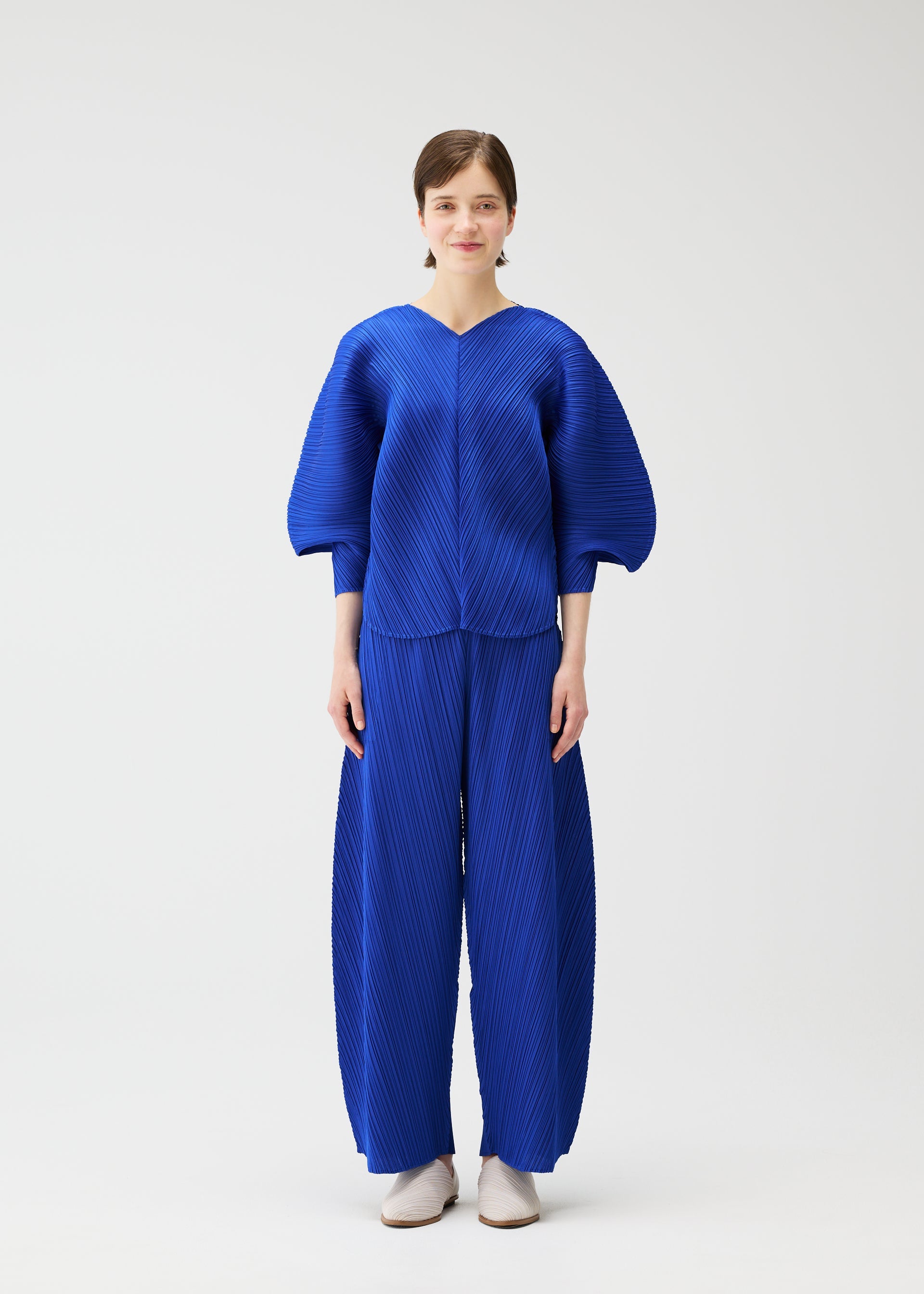 Zoomed view A woman with short brown hair and white shoes models the COAST set by PLEATS PLEASE ISSEY MIYAKE: a loose, bright blue top with a wave pattern and balloon sleeves, paired with matching wide-leg pants, all against a plain white background.